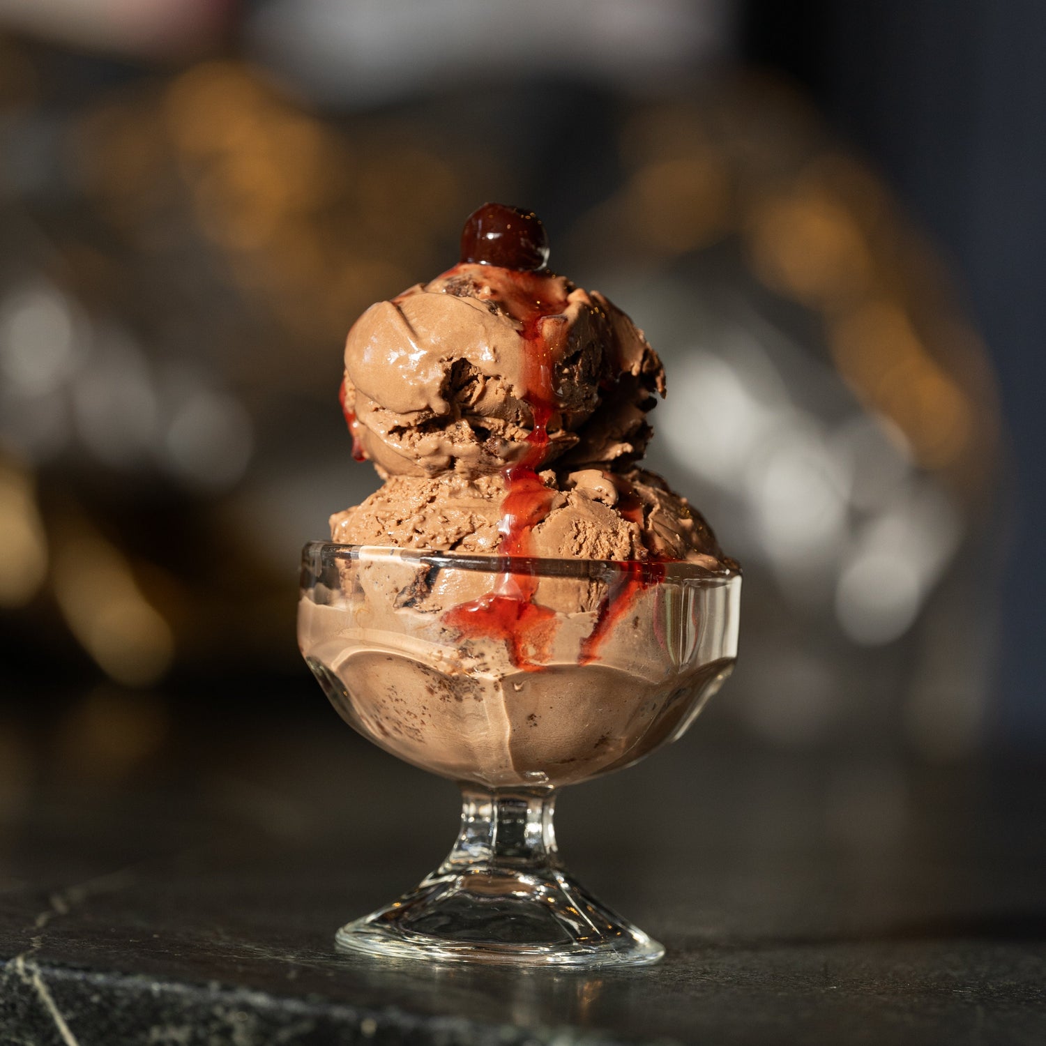 Two scoops of chocolate ice cream in a glass dish on a dark surface with a blurred background.
